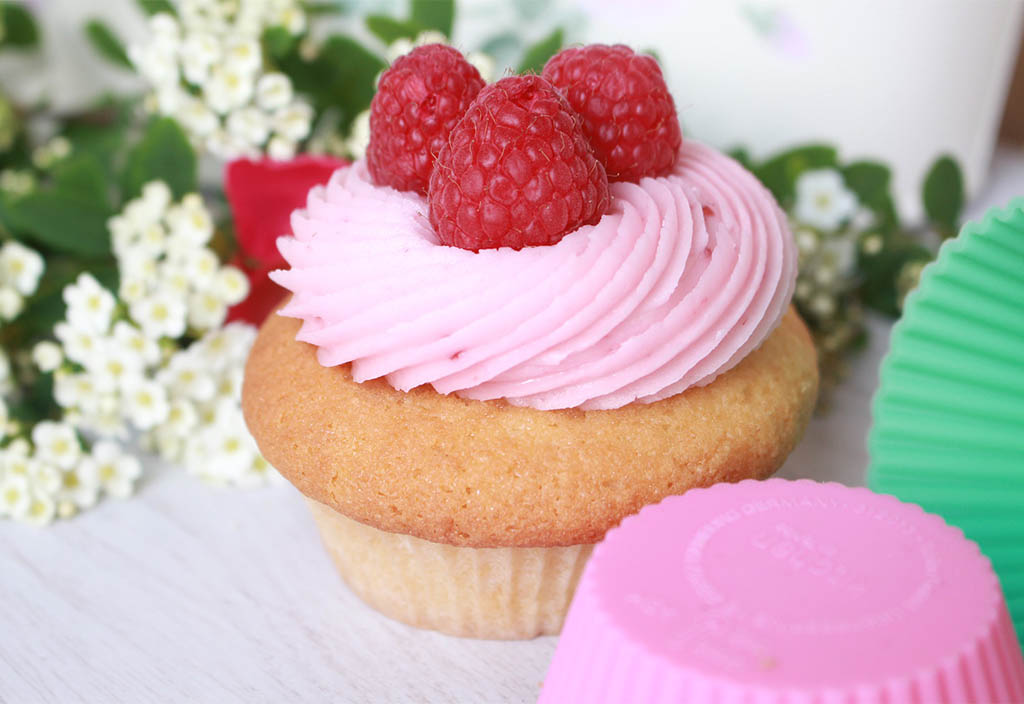 Vaniljmuffins dekorerad med rosa hallonsmörkräm och garnerad med hallon står bakom en rosa och grön muffinsform. I bakgrunden ligger vita blommor.