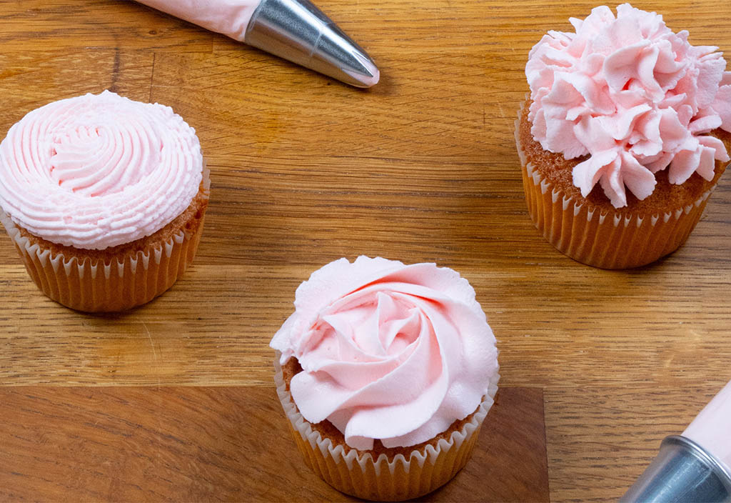 Tre vaniljmuffins med rosa smörkräm som garnering samt två spritsar innehållandes rosa smörkräm.