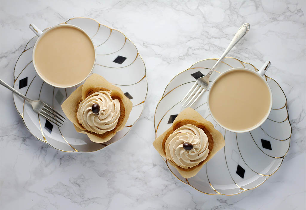 Två kaffekoppar innehållandes kaffe står på två assietter där även muffins täckta med mockasmörkräm garnerad med chokladknapp står. På tallrikarna ligger det även vars en silvrig gaffel.