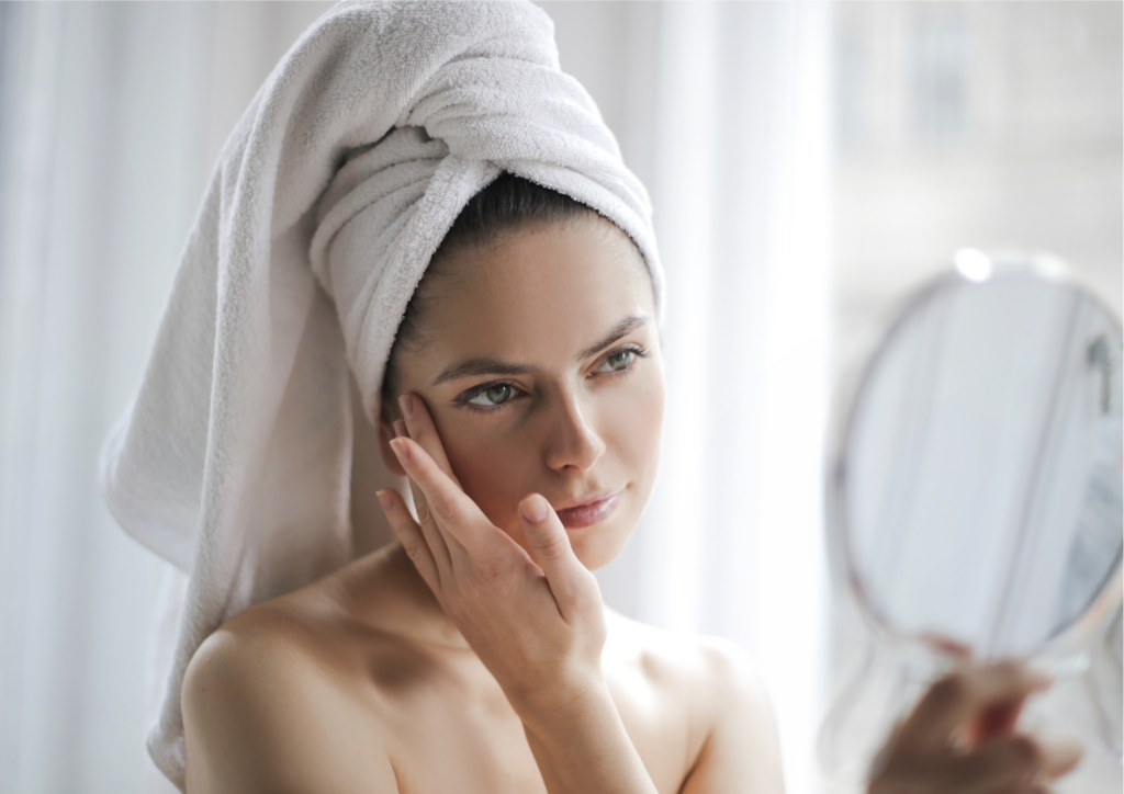 En kvinna som har handduk på huvudet samtidigt som hon tittar sig i spegeln och rör vid sin hud i ansiktet.