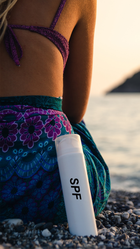 Kvinna sitter på den strand i bikini och sarong med en flaska lutandes mot sig med texten SPF. 