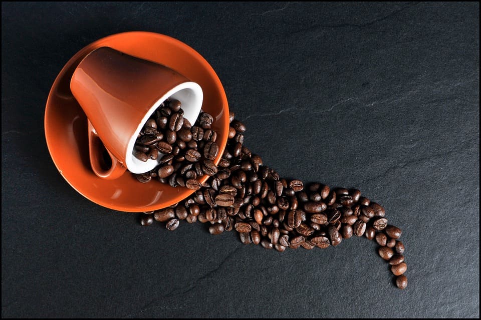En orange kaffekopp liggandes på sitt fat som spiller ut kaffebönor