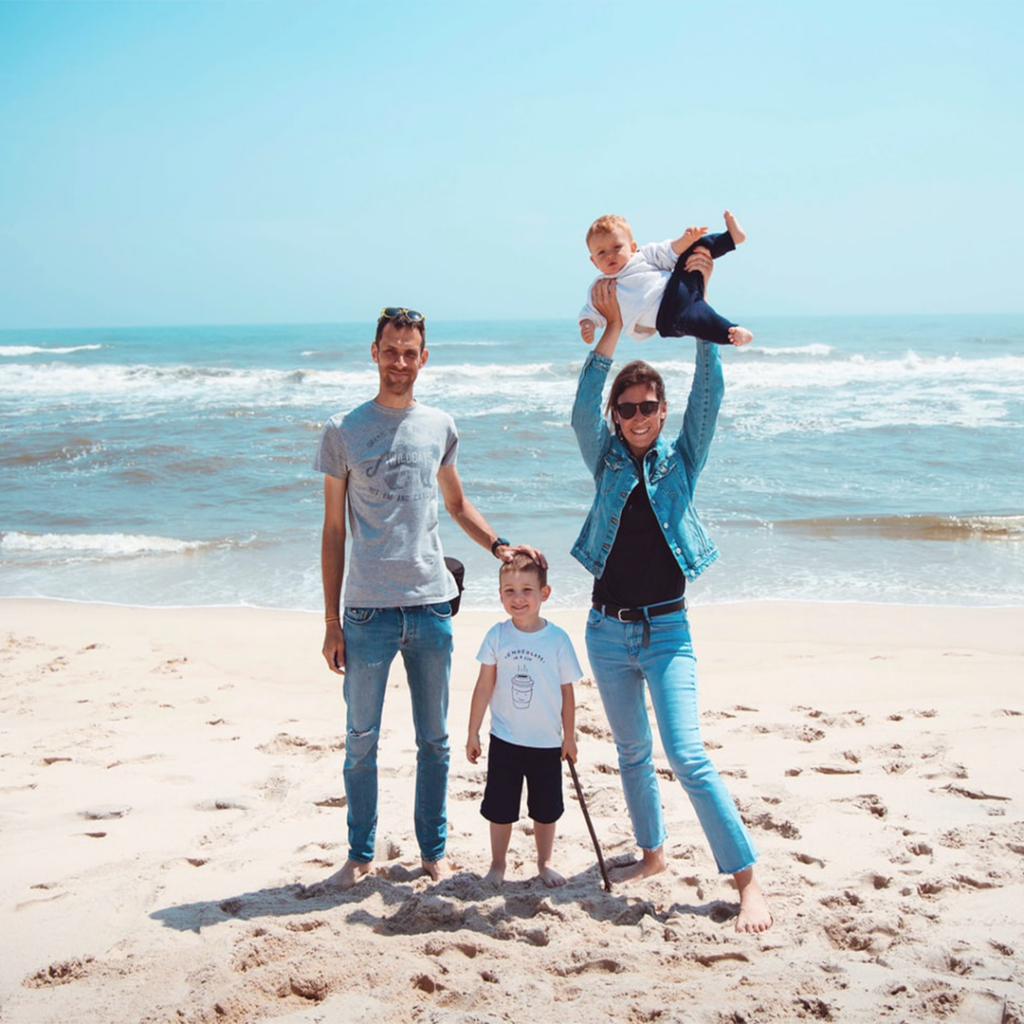En familj med två vuxna och två barn som står på stranden med havet bakom sig.