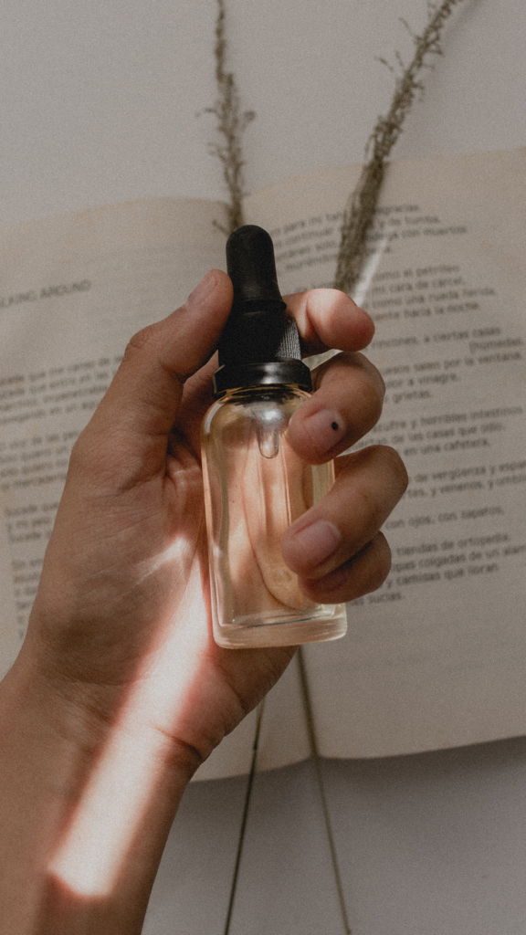En hand som håller i en flaska innehållande ett serum eller olja med pipett. Under ligger en öppen bok och två halmstrån.